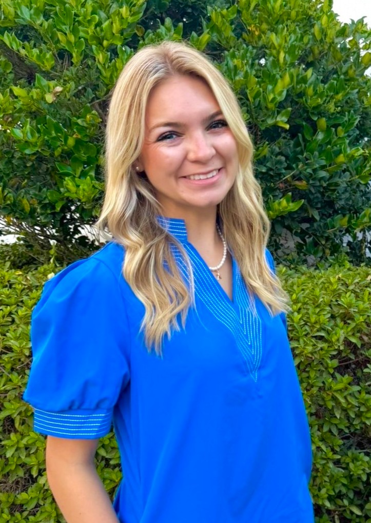 A smiling woman with blonde hair stands outdoors, wearing a vibrant blue blouse in front of green foliage.